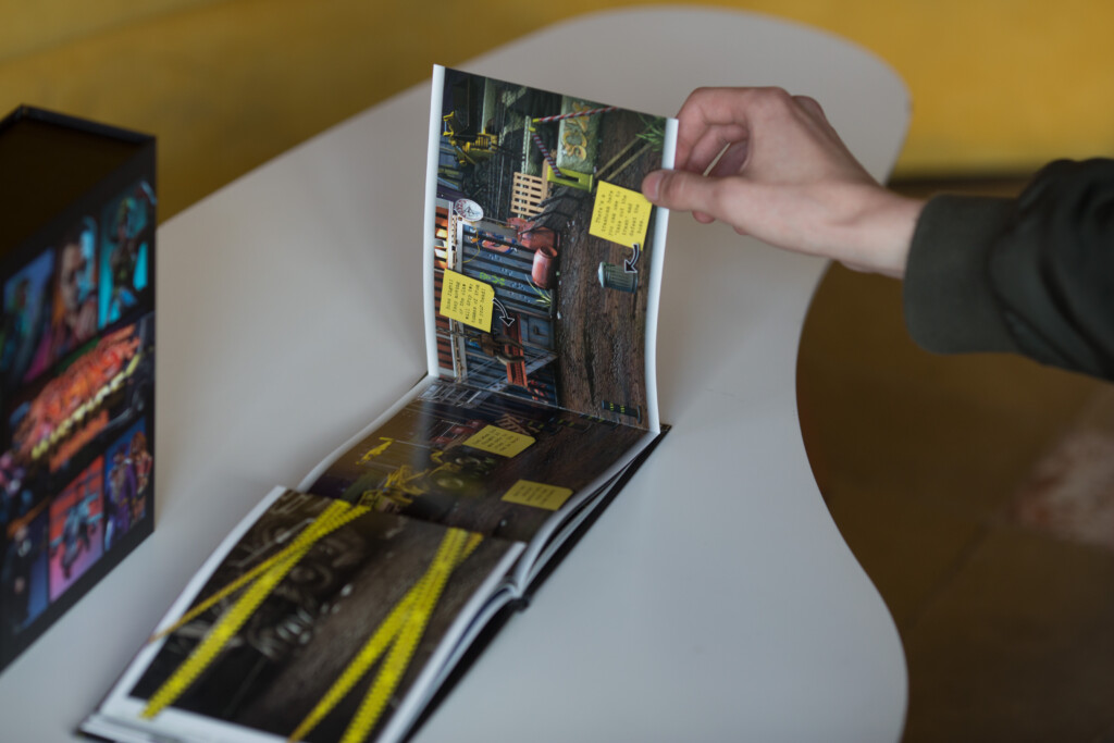 Hand holding open a hardcover book with yellow sticky notes and urban imagery visible on pages, displayed on white surface