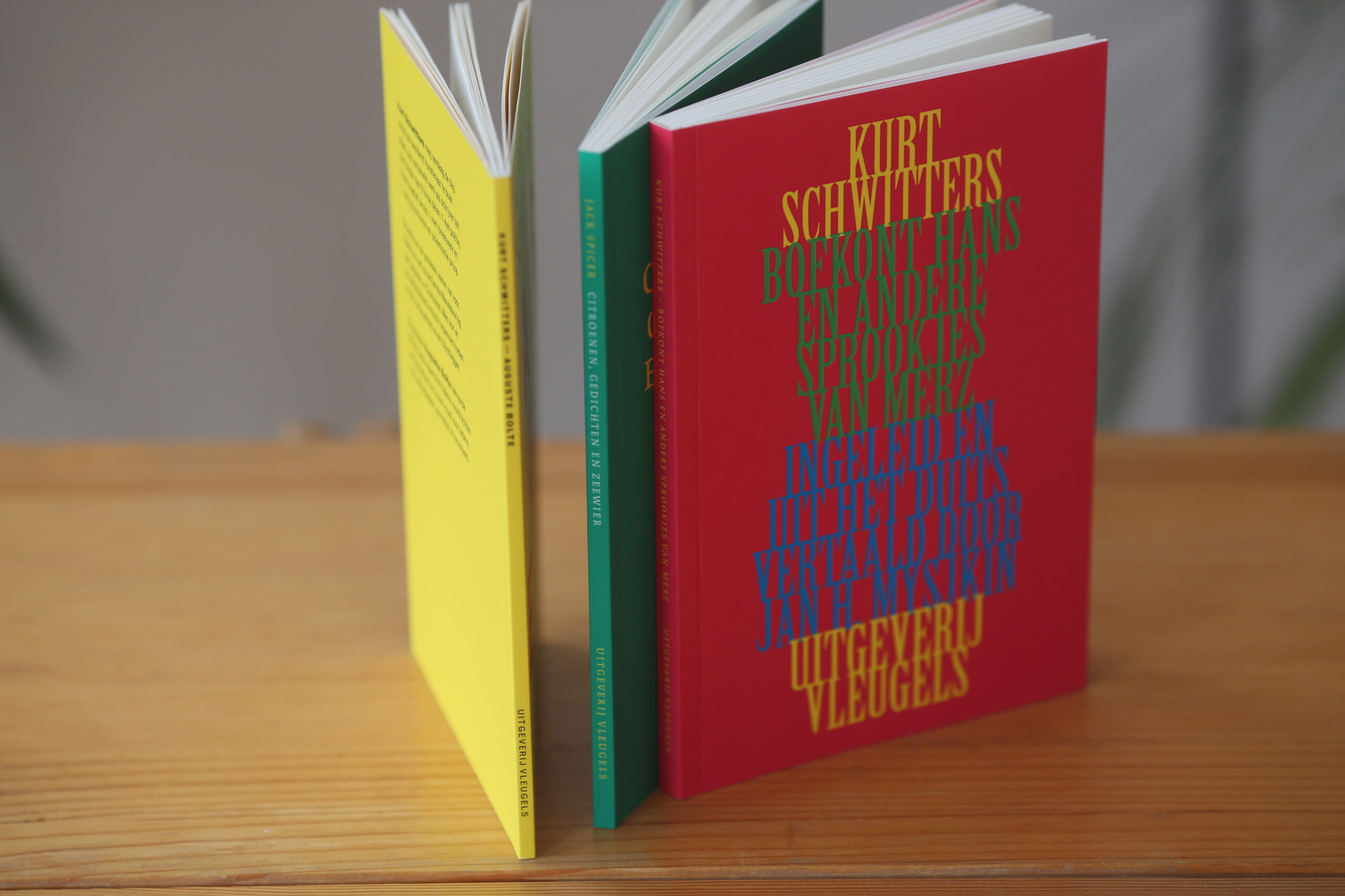 Three softcover books with yellow, green, and red covers fanned open on a wooden surface, the red cover printed with gold