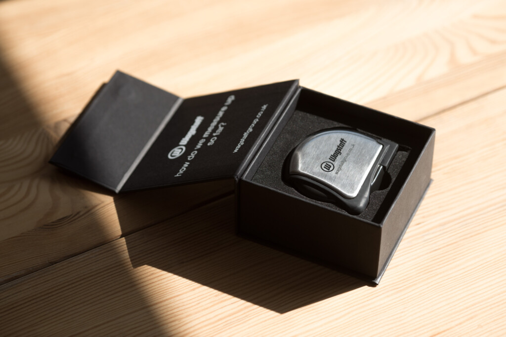 Black rigid box open to reveal grey product inside, displayed on light wood surface with sunlight casting shadows.