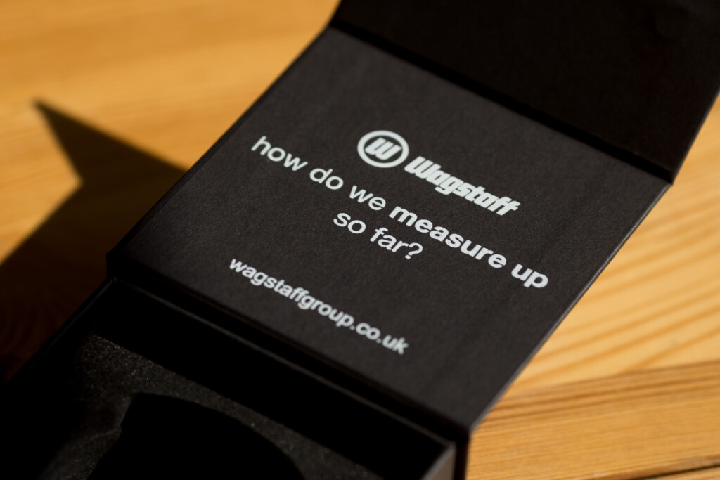 Black rigid box with white text reading "how do we measure up so far?" and Wagstaff logo, displayed on wooden surface.
