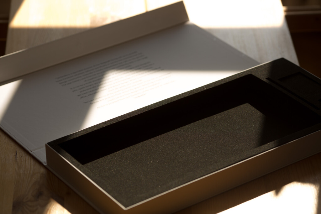 Open rigid box with charcoal grey interior and printed document visible behind it in natural light.