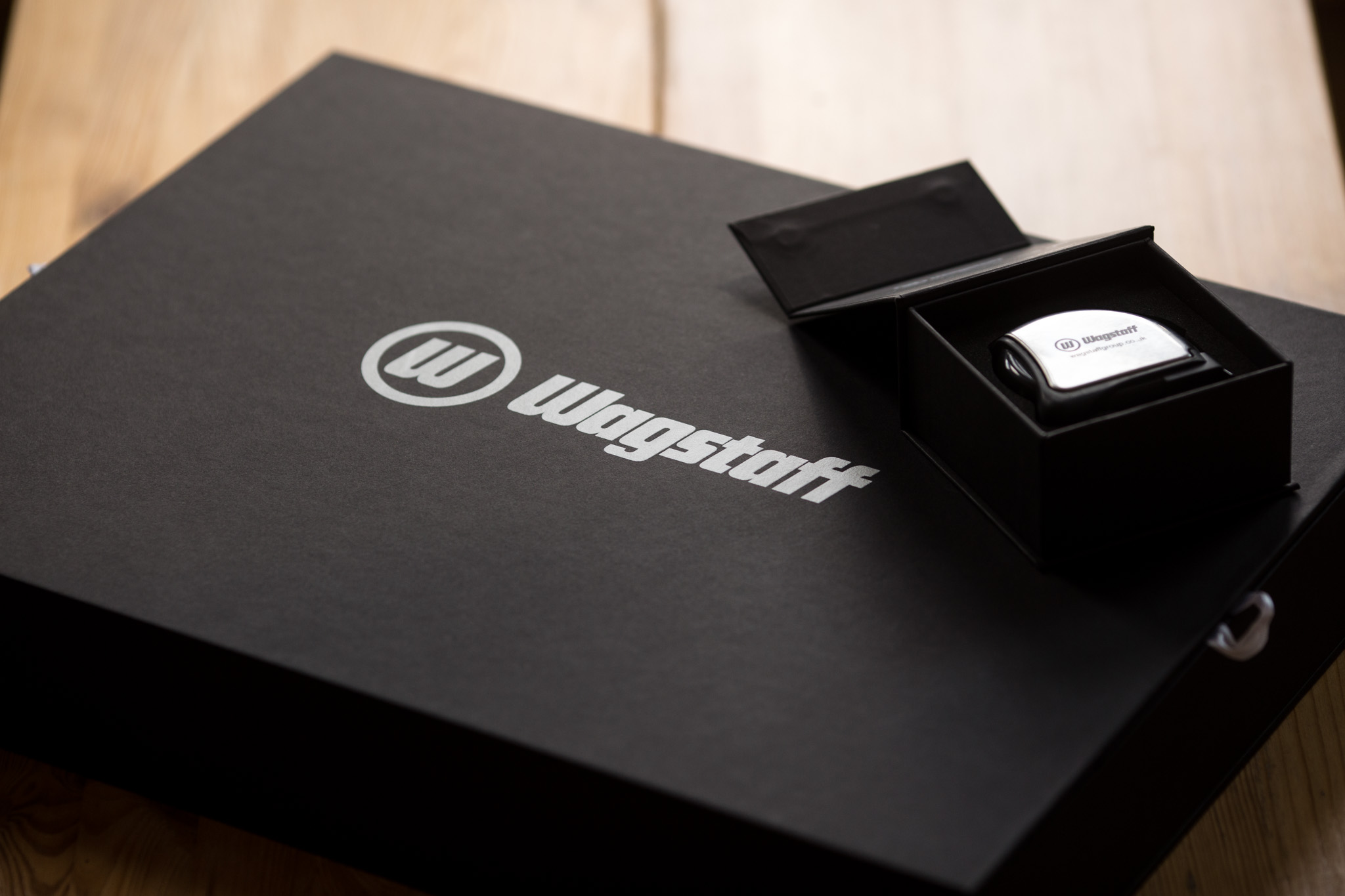 Black rigid boxes with white Wagstaff logo, one open to reveal a white business card inside, on wooden surface.