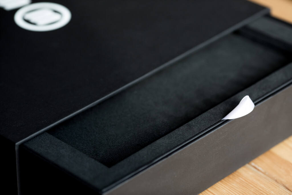 Black rigid box with foam insert and white ribbon detail, open to show interior packaging.