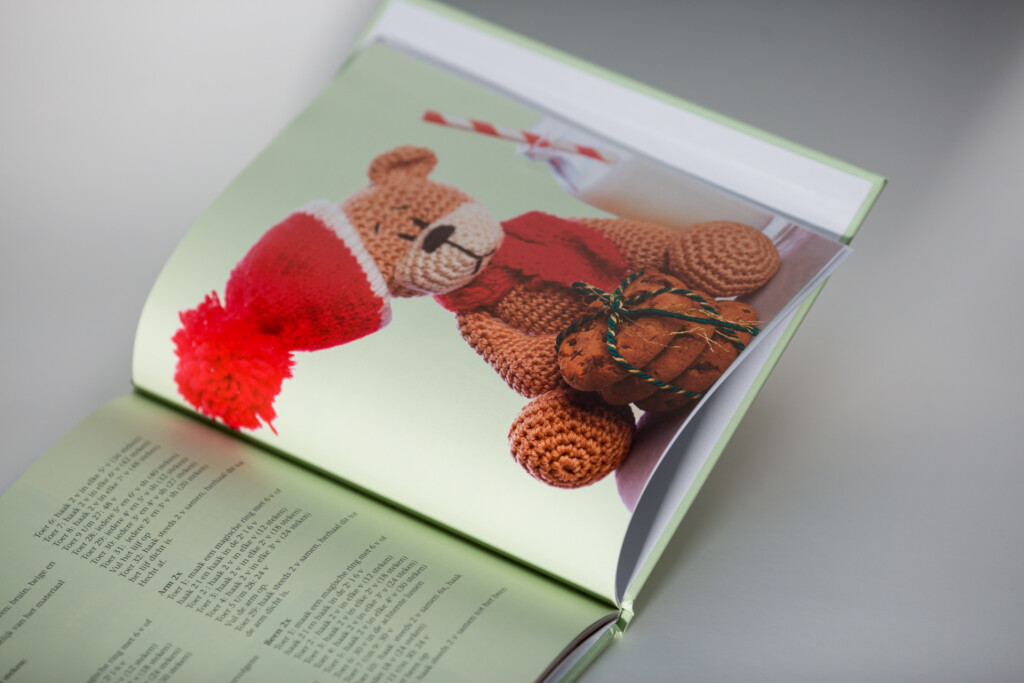 Hardcover book opened to show an interior spread with a crocheted teddy bear wearing a red bow tie and a red knitted hat