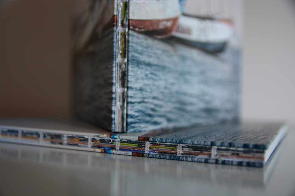 Spine detail of open hardcover book showing stitched binding and colourful endpapers, with stacked copies blurred in