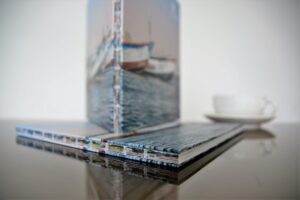 Stack of open spine hardcover books with water and boat imagery on covers, displayed on reflective surface with coffee cup.