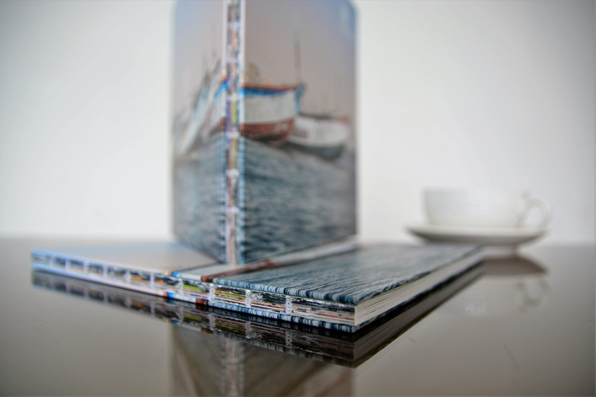 Stack of open spine hardcover books with water and boat imagery on covers, displayed on reflective surface with coffee cup.