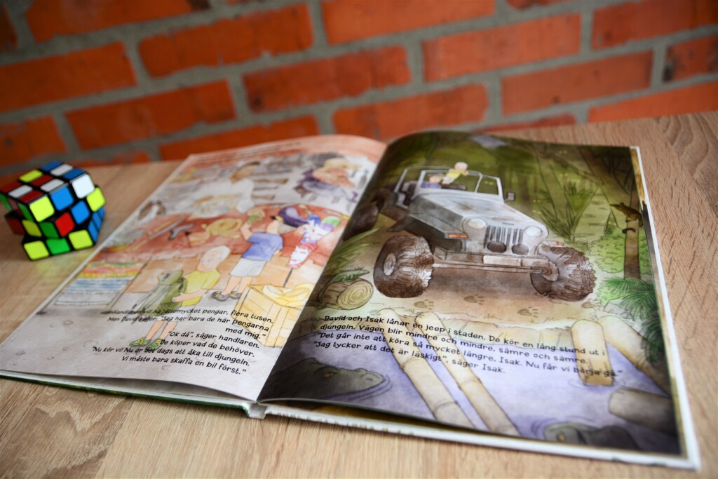 Open hardcover children's book showing illustrated page of a monster truck with text, beside a Rubik's cube on wooden