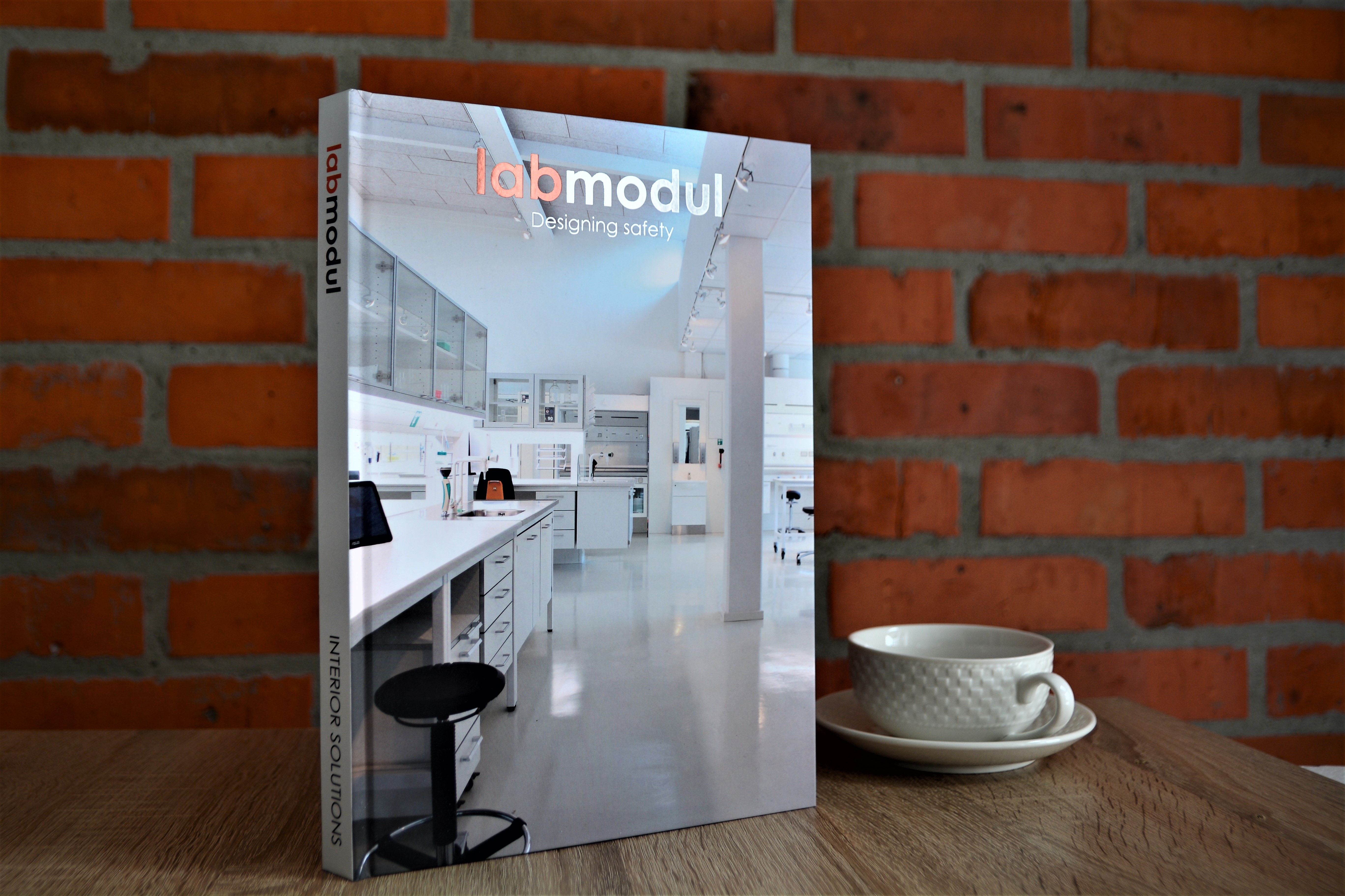Hardcover catalogue with hot stamping featuring two different foils, displayed upright against a brick wall with a coffee