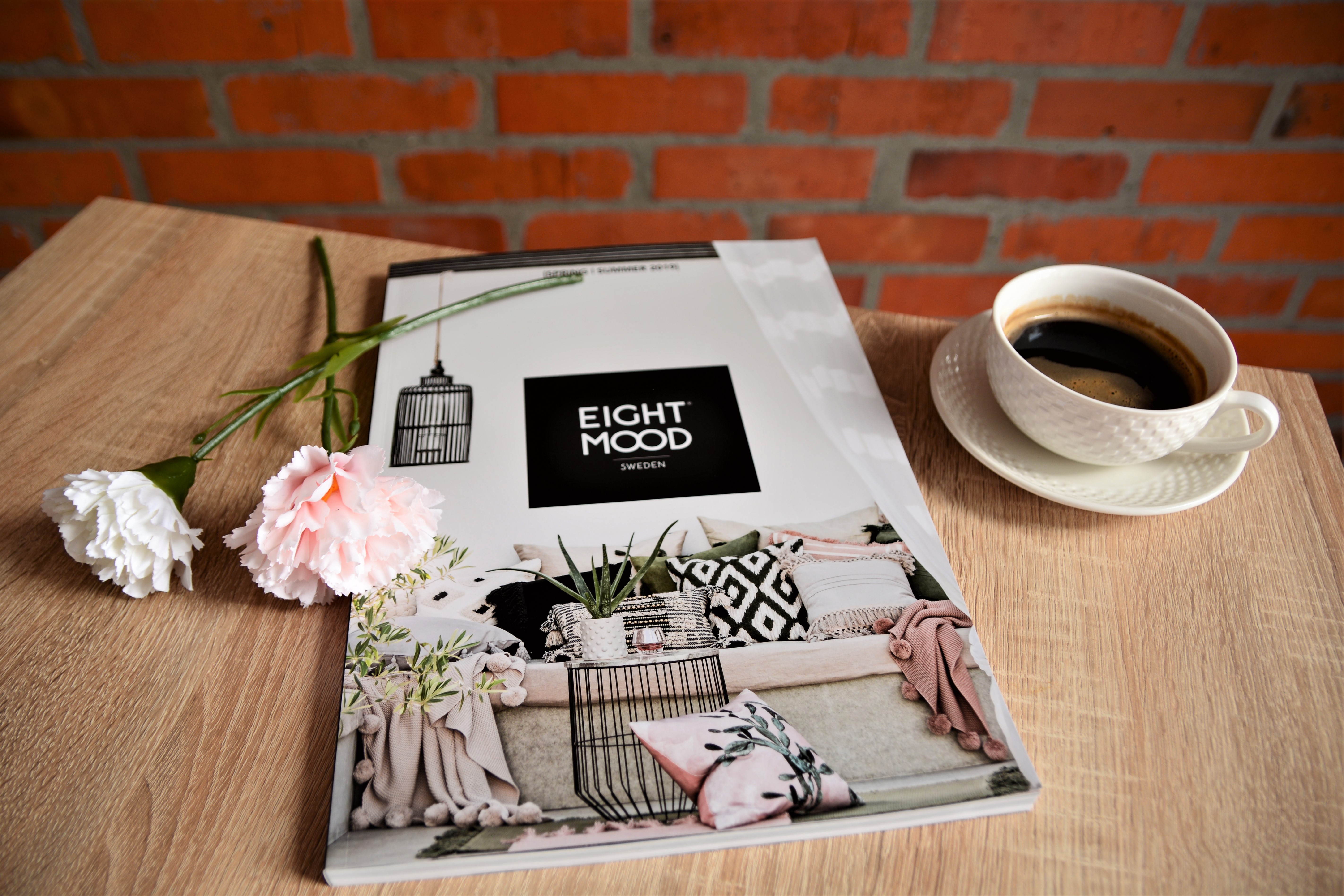 Printed catalogue with black label reading "EIGHT HOOD" on cover, displayed on wooden table with pink and white carnations