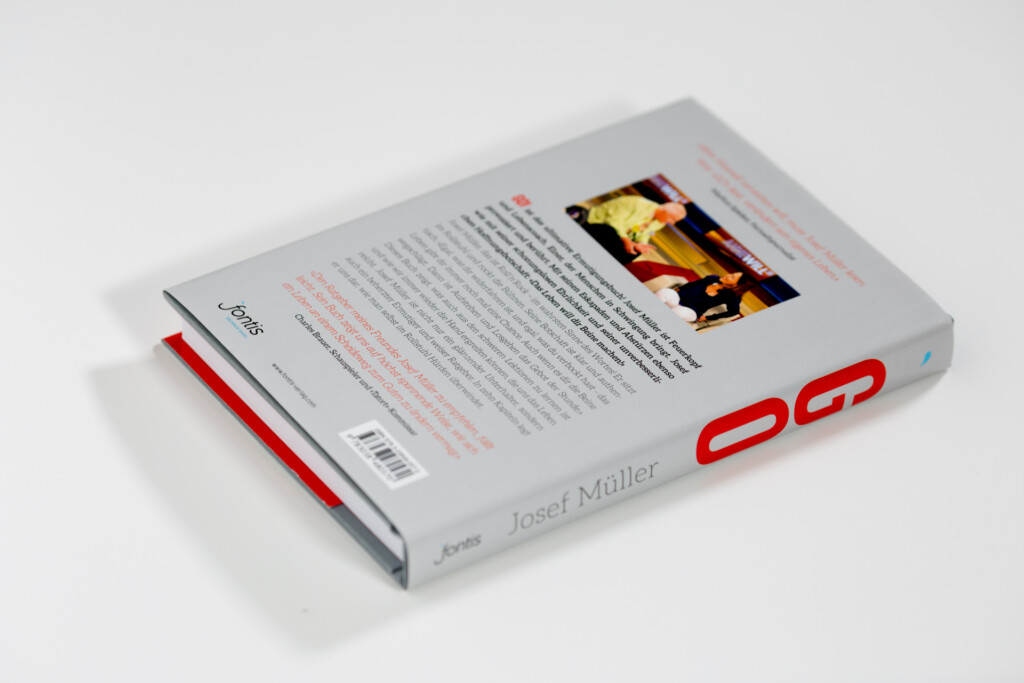 Hardcover book with grey wrapper and red spine detail, opened to show interior text and photograph, displayed against white