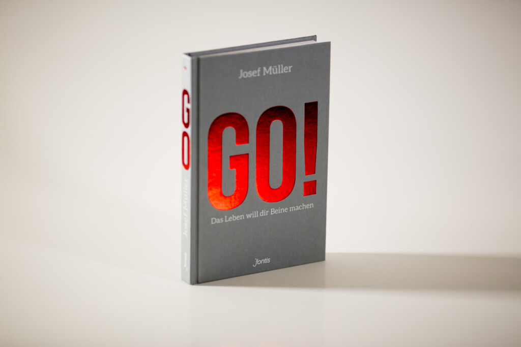 Hardcover book with grey cover and red foil-stamped title "GO!" by Josef Müller, displayed at an angle on a white surface.