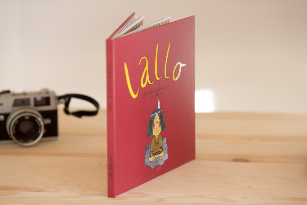 Red hardcover children's book titled "tallo" with illustrated bottle graphic on cover, displayed with vintage camera on