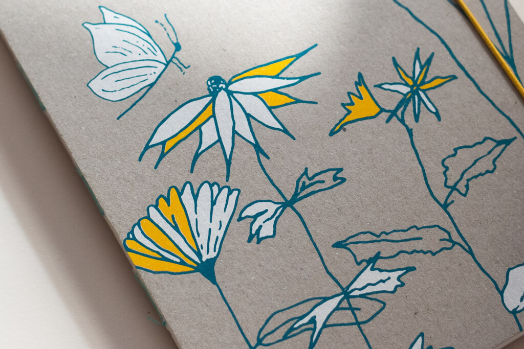 Printed notebook cover featuring hand-drawn botanical illustrations with blue pen outlines and yellow watercolour accents on