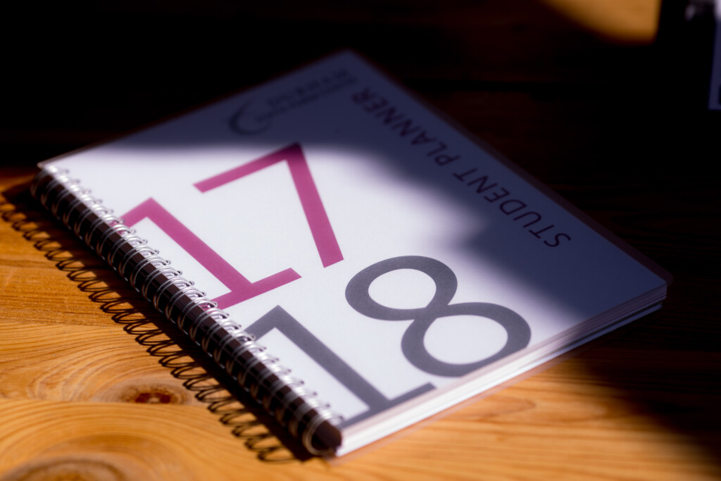 Spiral-bound student planner with purple and grey numbers 17 and 18 on white cover, lying on wooden surface.