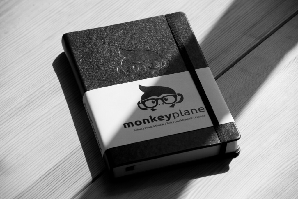 Black hardcover notebook with elastic band and embossed monkey logo wearing glasses on the cover, labelled monkeyplane.