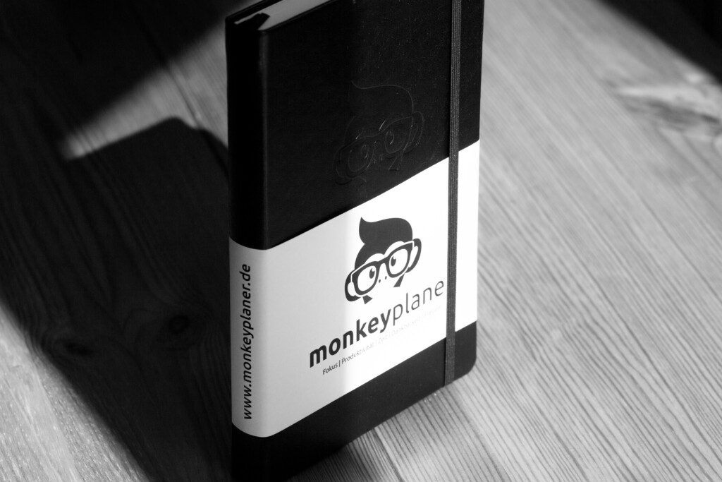Monkeyplane branded hardcover notebook with black cover and white label on wooden surface, black and white studio photograph.