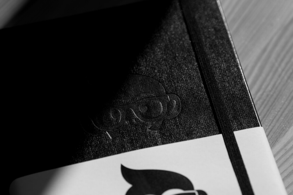 Black hardcover notebook with elastic band and embossed logo on cover, shown at angle on lined surface.