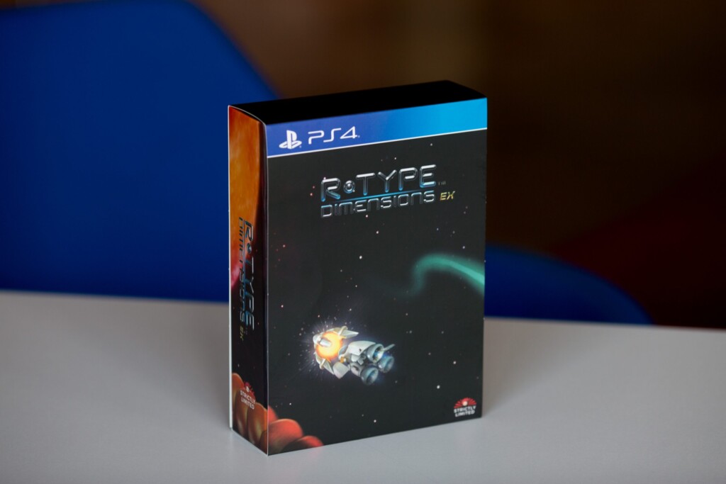 R-Type Deluxers EX PS4 game box with blue stripe, black cover featuring spacecraft illustration, displayed against blurred