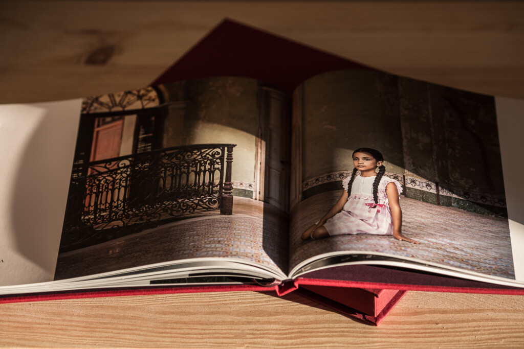 Open hardcover photo book displaying interior pages with ornate balcony on left and young girl in white dress on right, with