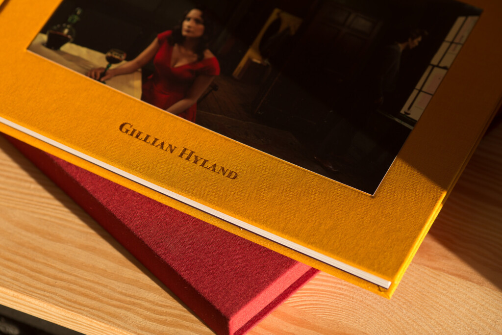 Hardcover photo album with gold cloth cover stamped with "Gillian Hyland", red ribbon bookmark, displayed open on wooden
