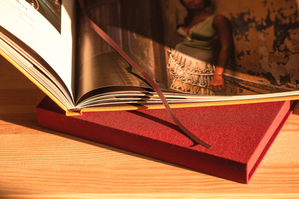 Open hardcover book with red cloth cover and ribbon bookmark on wooden surface, displaying printed pages with photographs.