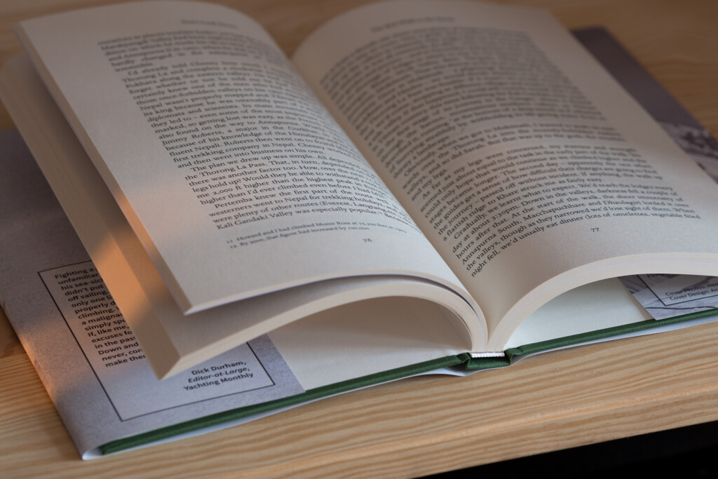 Hardcover book open to pages 76 and 77 showing dense text columns, displayed on a wooden surface with afternoon sunlight