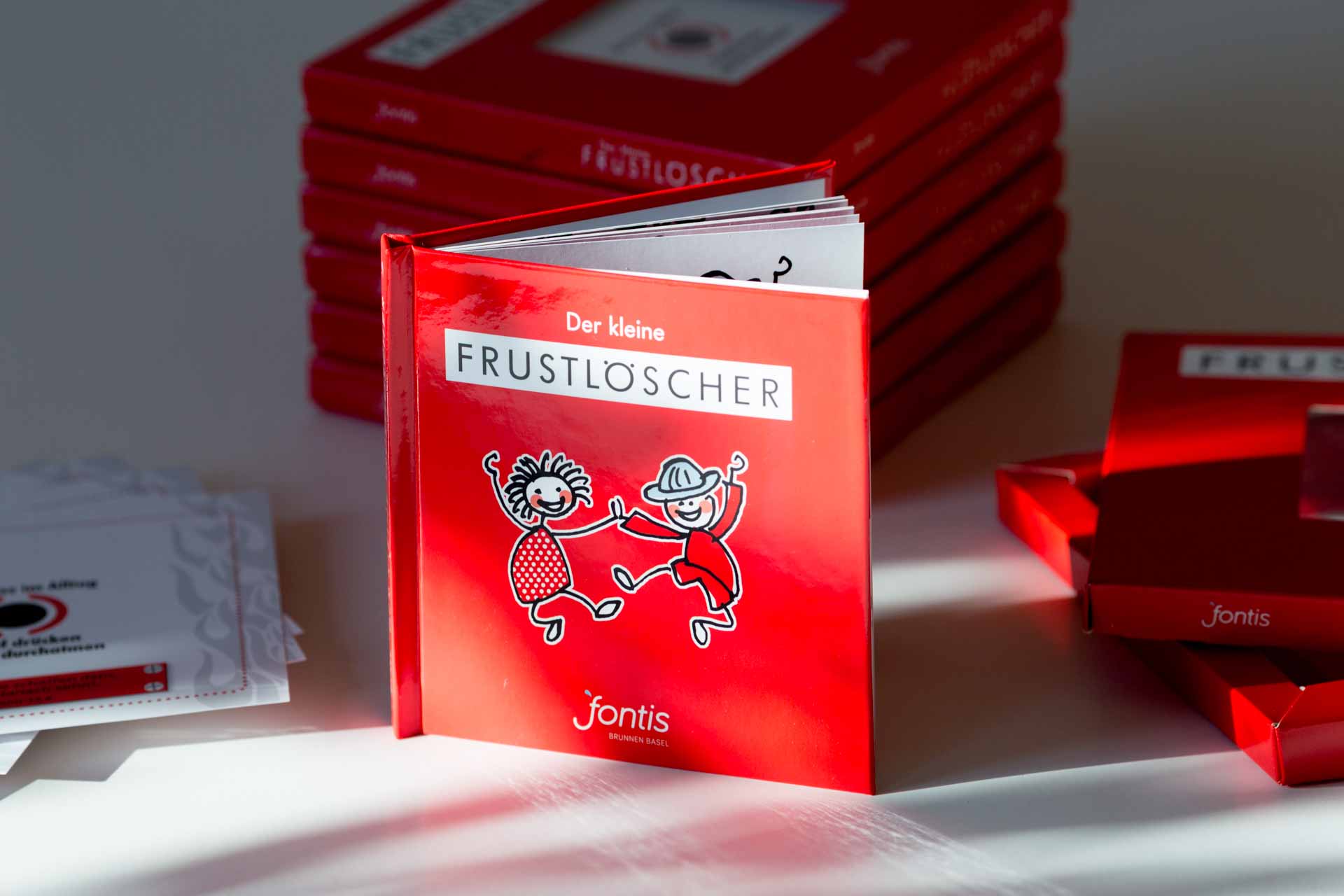 Red hardcover children's book titled Der kleine FRUSTLOSCHER with illustration of two figures, stacked copies visible in