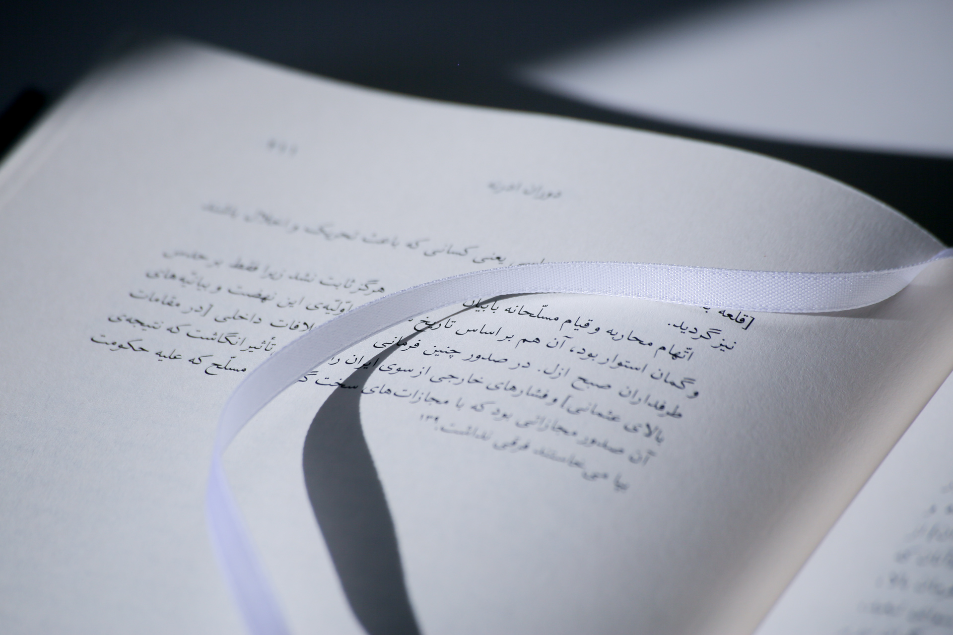Open hardcover book with white ribbon bookmark, pages showing text in Arabic script.