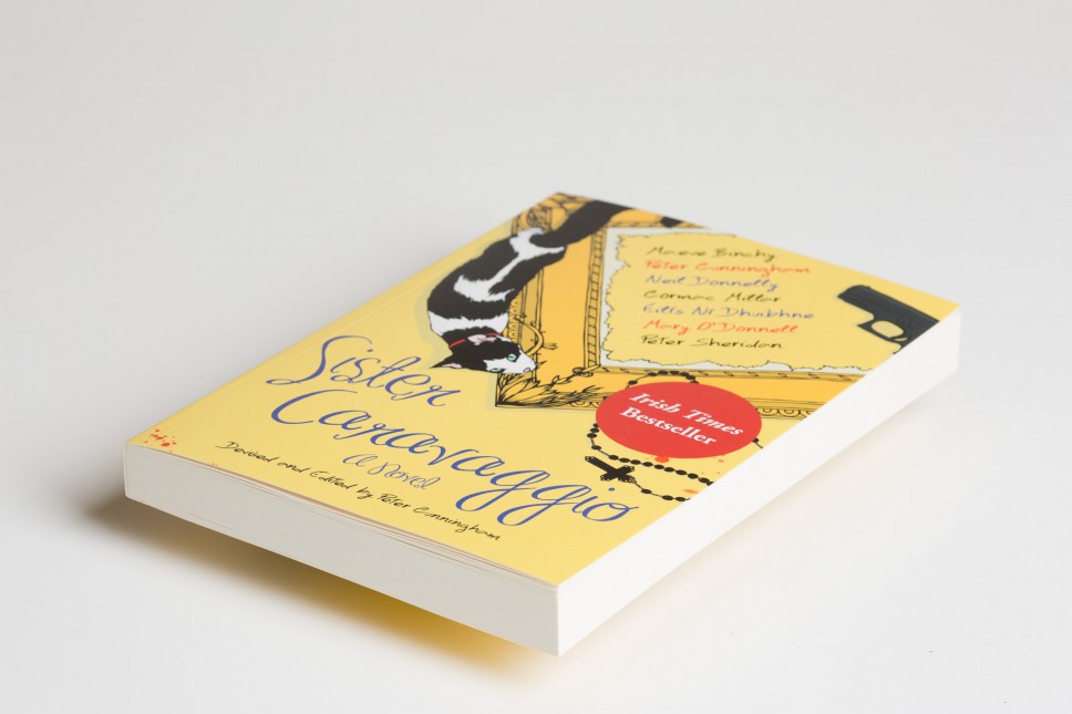 Softcover book with yellow cover featuring illustrated figures and handwritten text, displayed at an angle against a plain