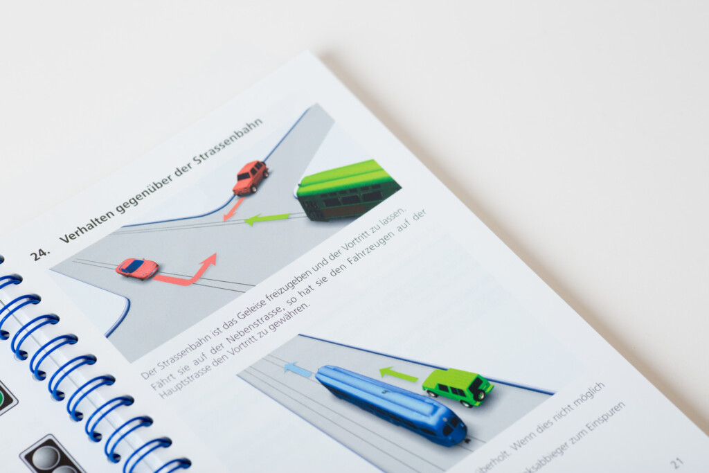 Open spiral-bound book showing page 24 with diagrams of coloured toy cars on track layouts and a paintbrush demonstration.