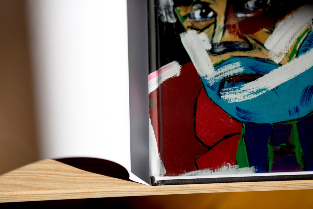 Hardcover art book with colourful abstract portrait on cover, displayed open on wooden surface showing interior pages.