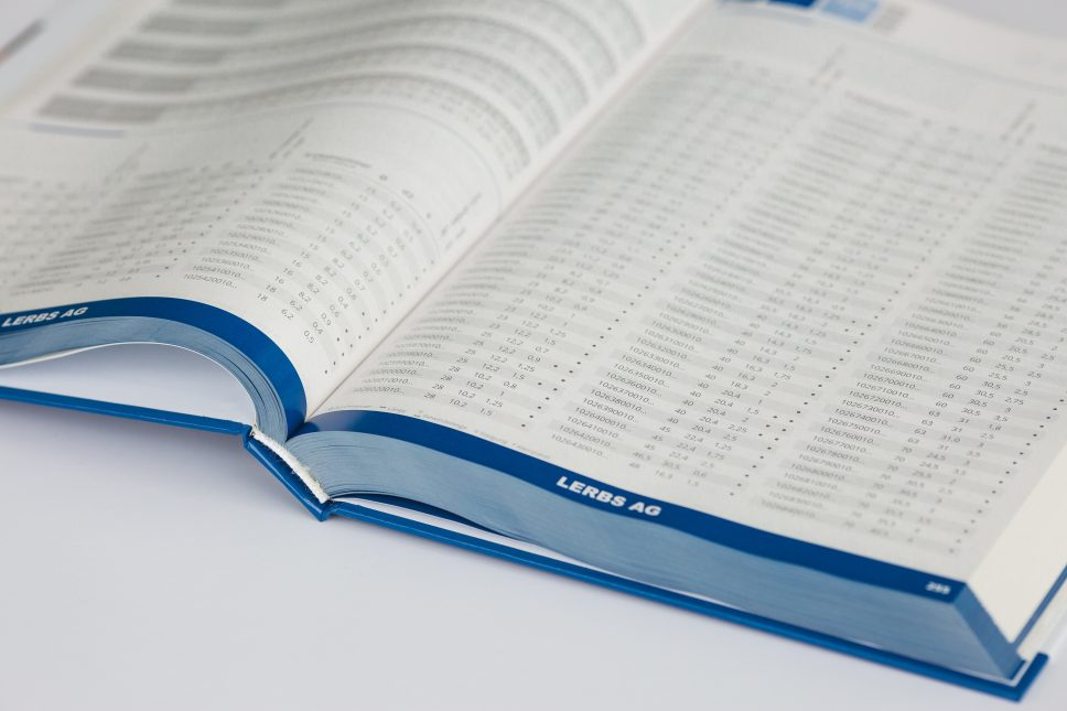 Open hardcover catalogue with blue ribbon bookmark, showing pages of data tables and text.