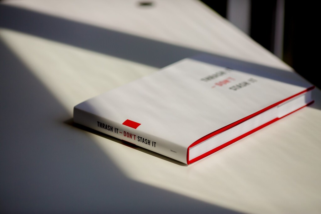 Hardcover catalogue with red edges lying on a white surface in sunlight, spine reading "Thrash it - Don't Stash it".