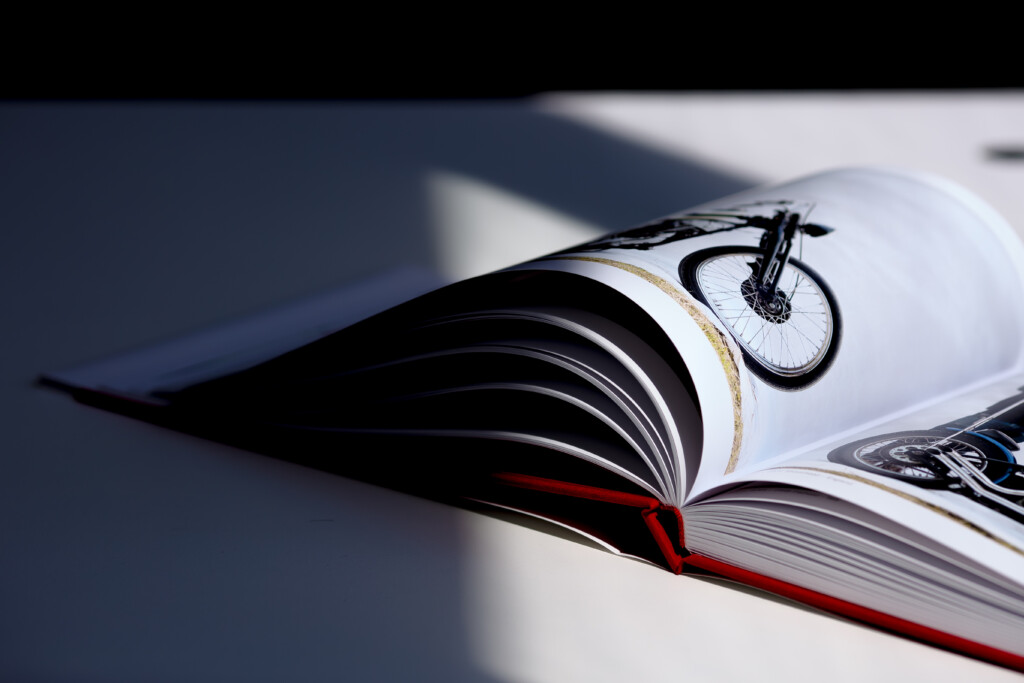 Open hardcover catalogue with pages fanned out, displaying a bicycle illustration on the visible spread with red ribbon