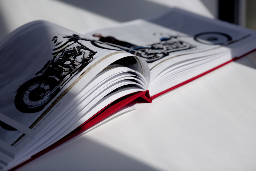 Open hardcover catalogue with red ribbon bookmark showing black and white motorcycle illustration on left page.