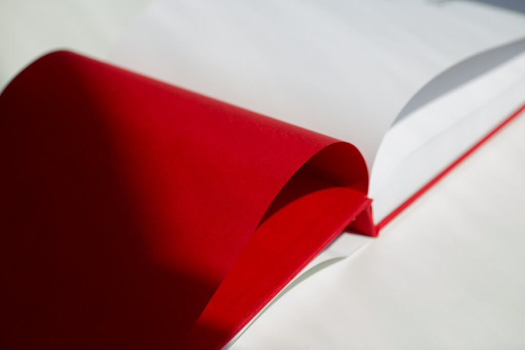 Red hardcover catalogue with white ribbon bookmark, fanned open to show binding detail.
