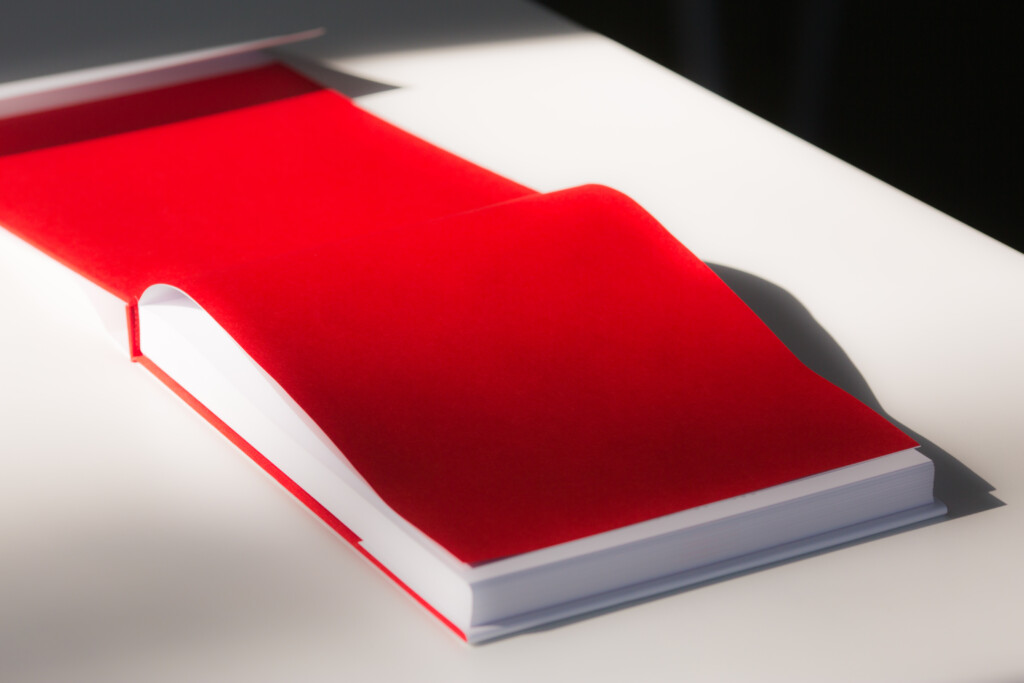 Red hardcover catalogue with white pages and grey binding, displayed open at an angle.