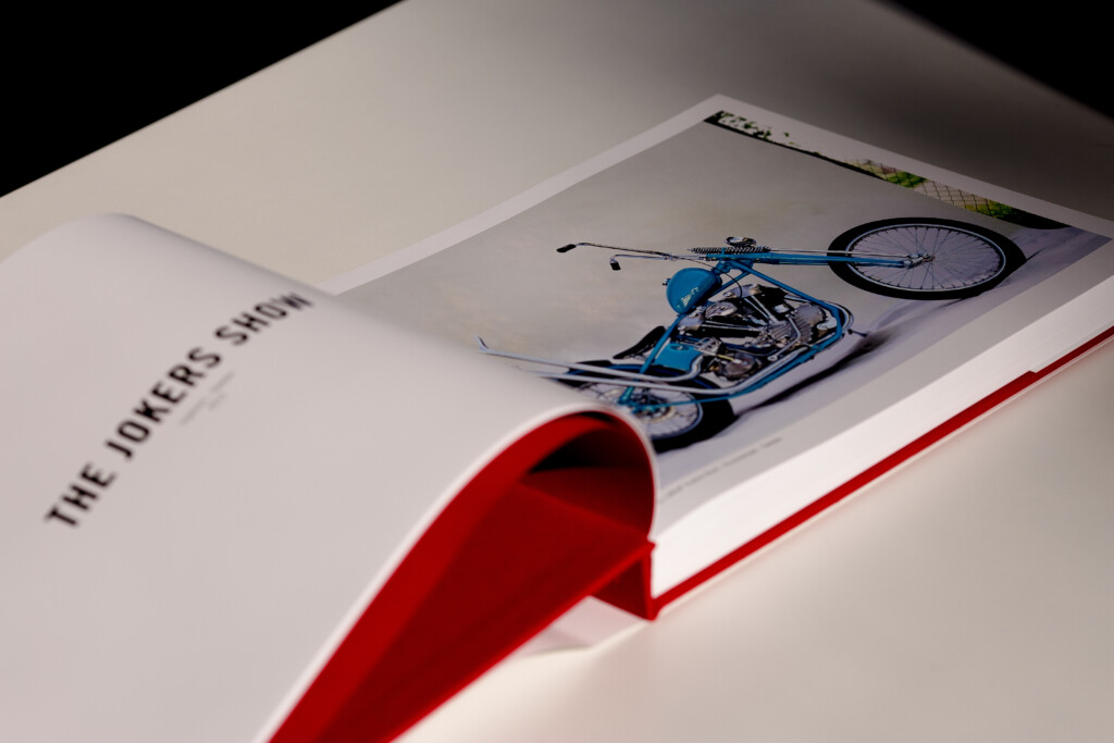Hardcover catalogue with red ribbon bookmark, open to show blue motorcycle illustration on right page and text on left page.