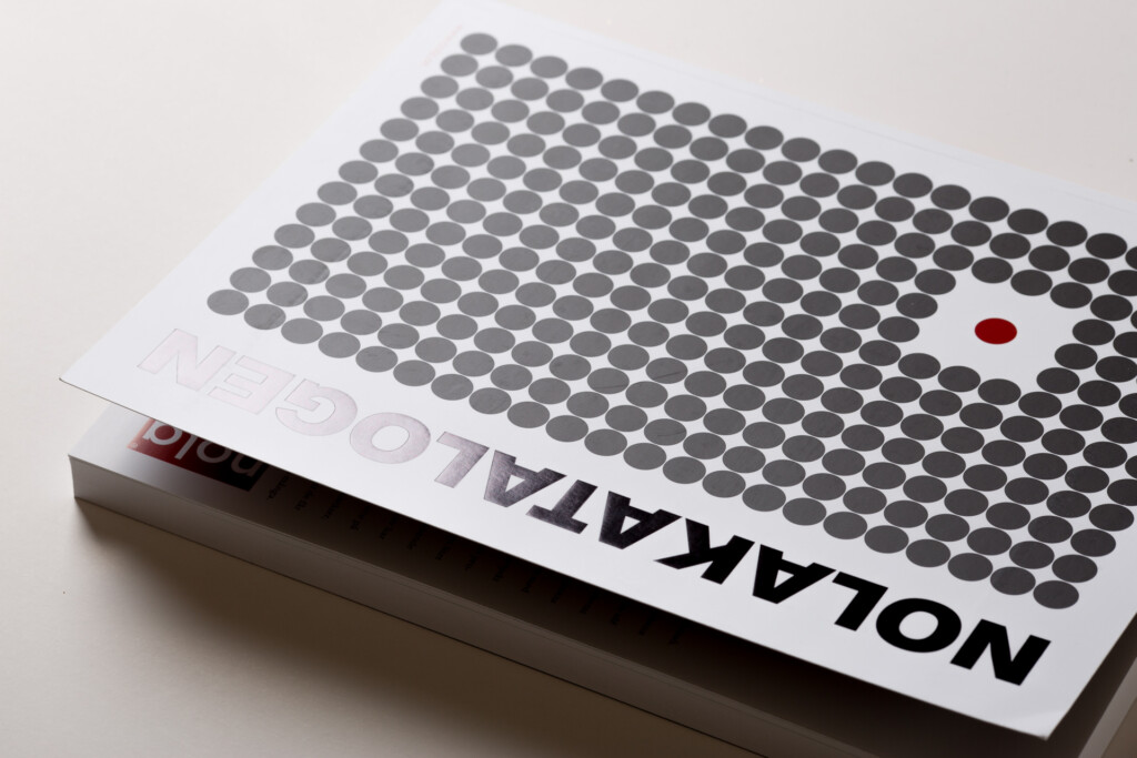 Nola product catalogue with grey circular dots arranged in a grid pattern and one red dot, printed on white paper.