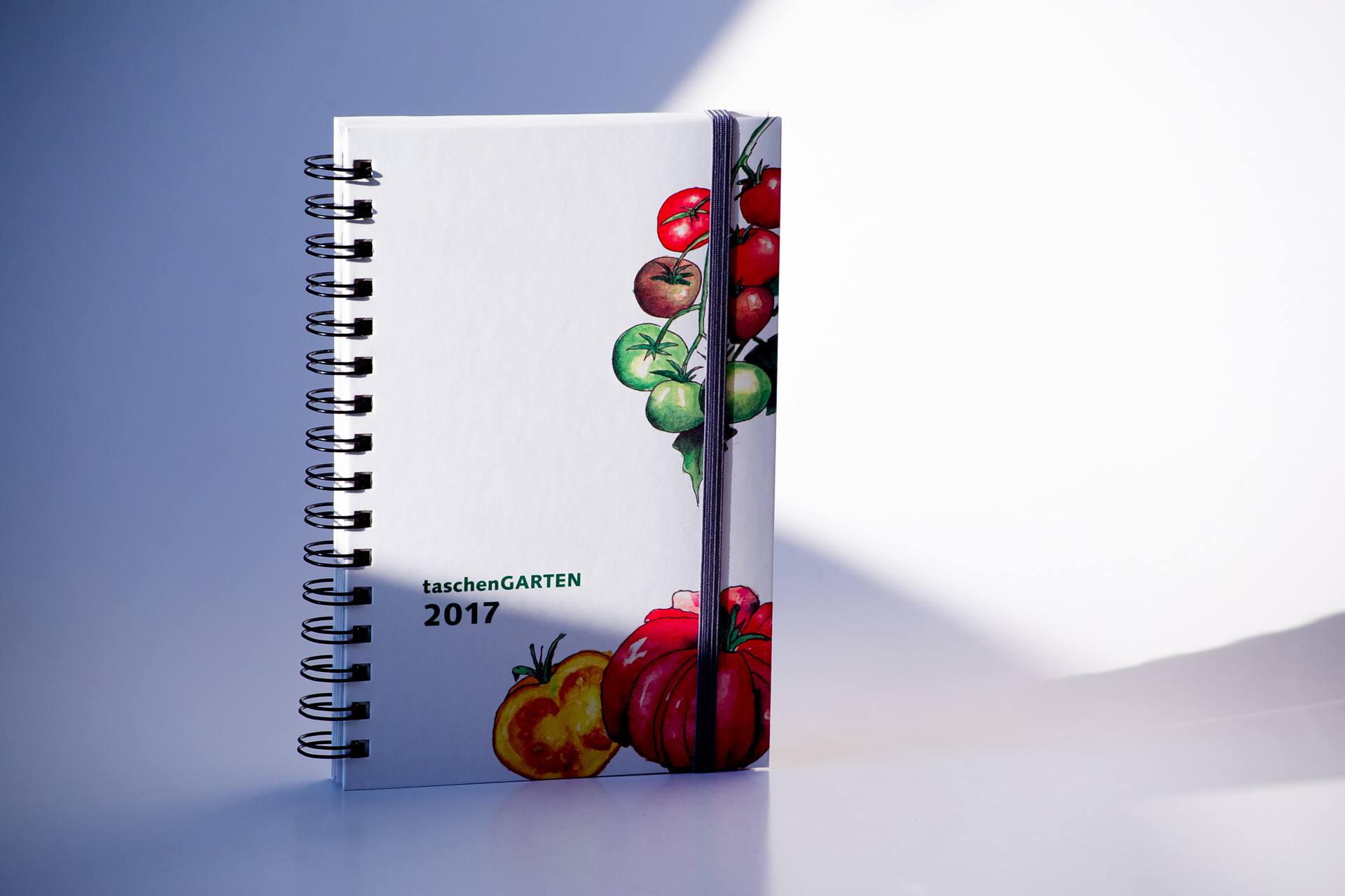 Spiral-bound notebook with illustrated cover featuring red and green tomatoes, titled taschenGARTEN 2017, shown with elastic