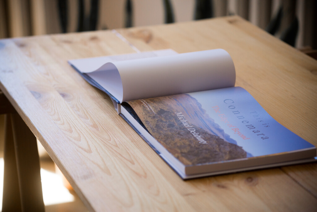 Rolled white paper and open softcover catalogue with blue cover and landscape photograph, displayed on wooden table.