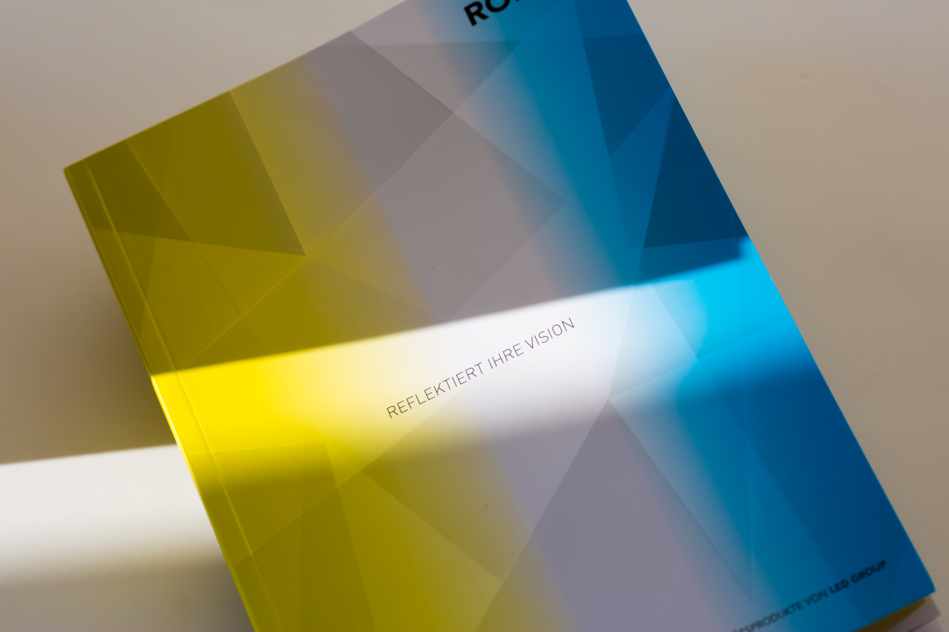 Printed catalogue with yellow and blue geometric covers fanned open, showing reflective finish and light beam across pages.
