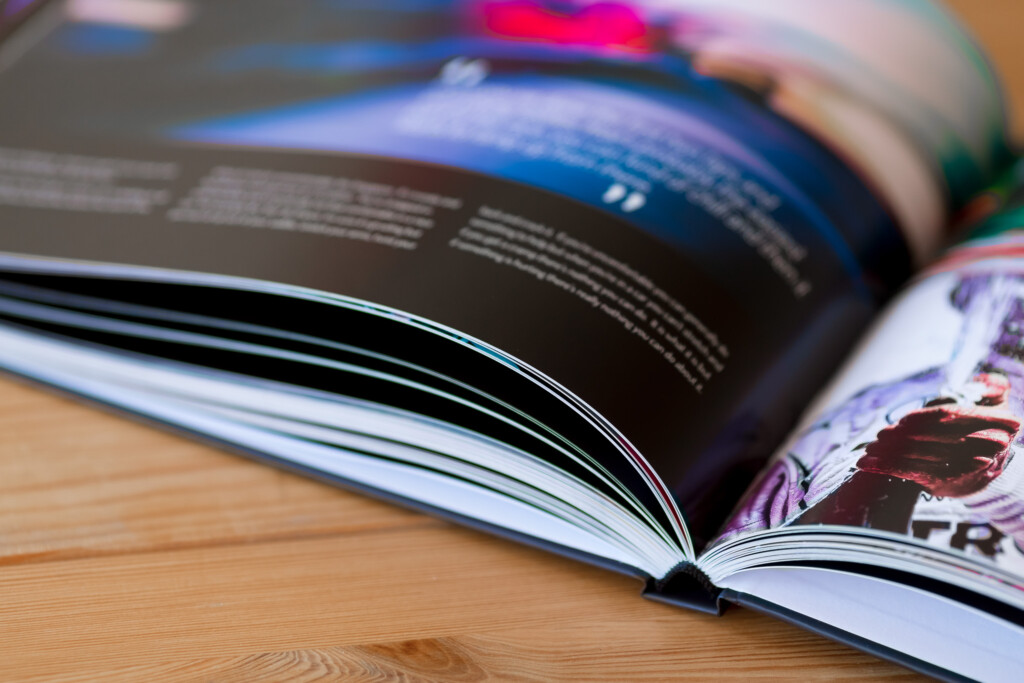 Open hardcover photo album with glossy pages fanned out on wooden surface, showing colour photography and text.