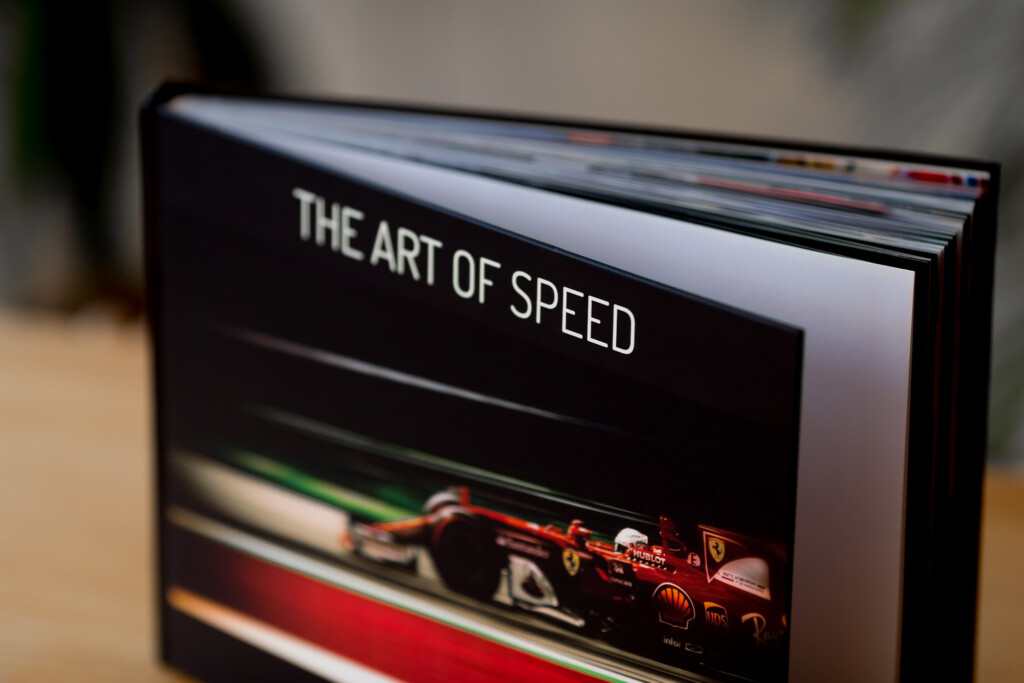 Hardcover book titled "The Art of Speed" with red Ferrari race car on cover, pages fanned open to show interior.