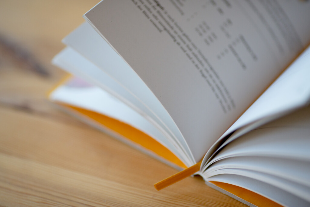 Open hardcover book with pages fanned out, showing text and images on the visible spread, with a yellow ribbon bookmark.