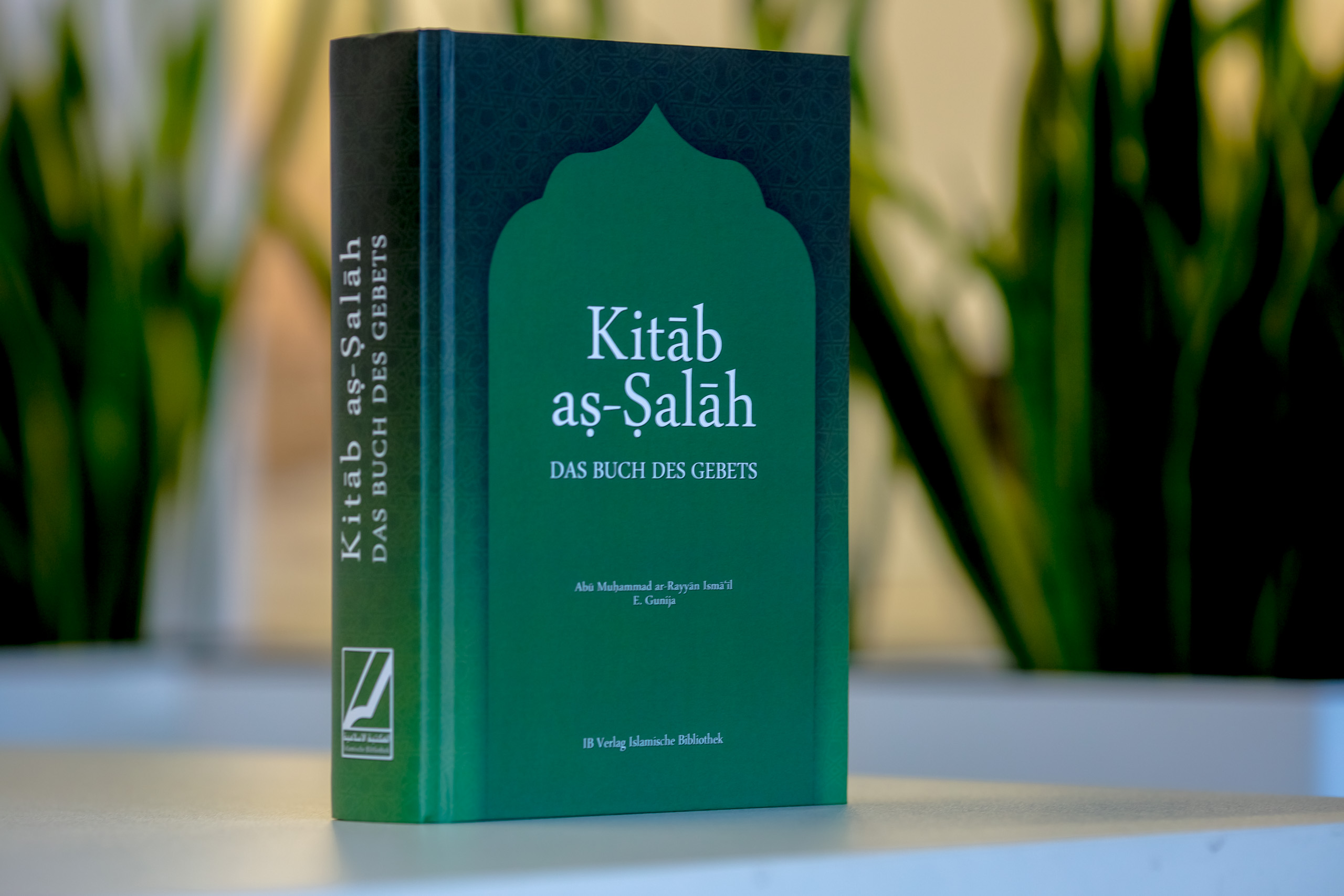 Hardcover book with dark teal cover featuring green Islamic arch design and "Kitab as-Salah" title, displayed outdoors with