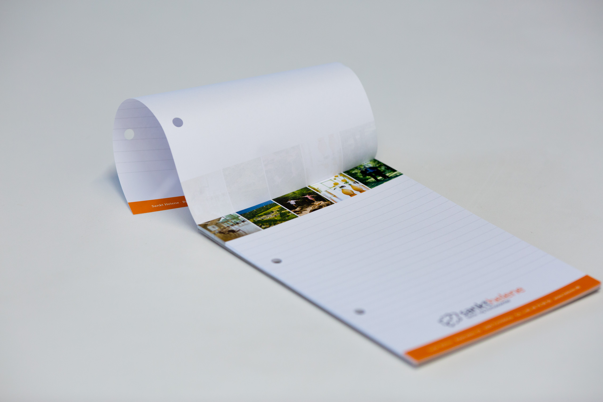 White spiral-bound notepad with orange accents, three landscape photographs visible on the binding strip, open to show blank