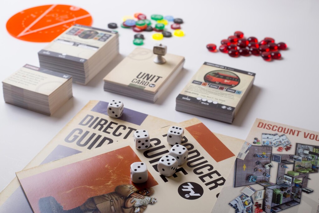Board game components including rulebooks, dice, coloured tokens, and red beads arranged on a white surface.