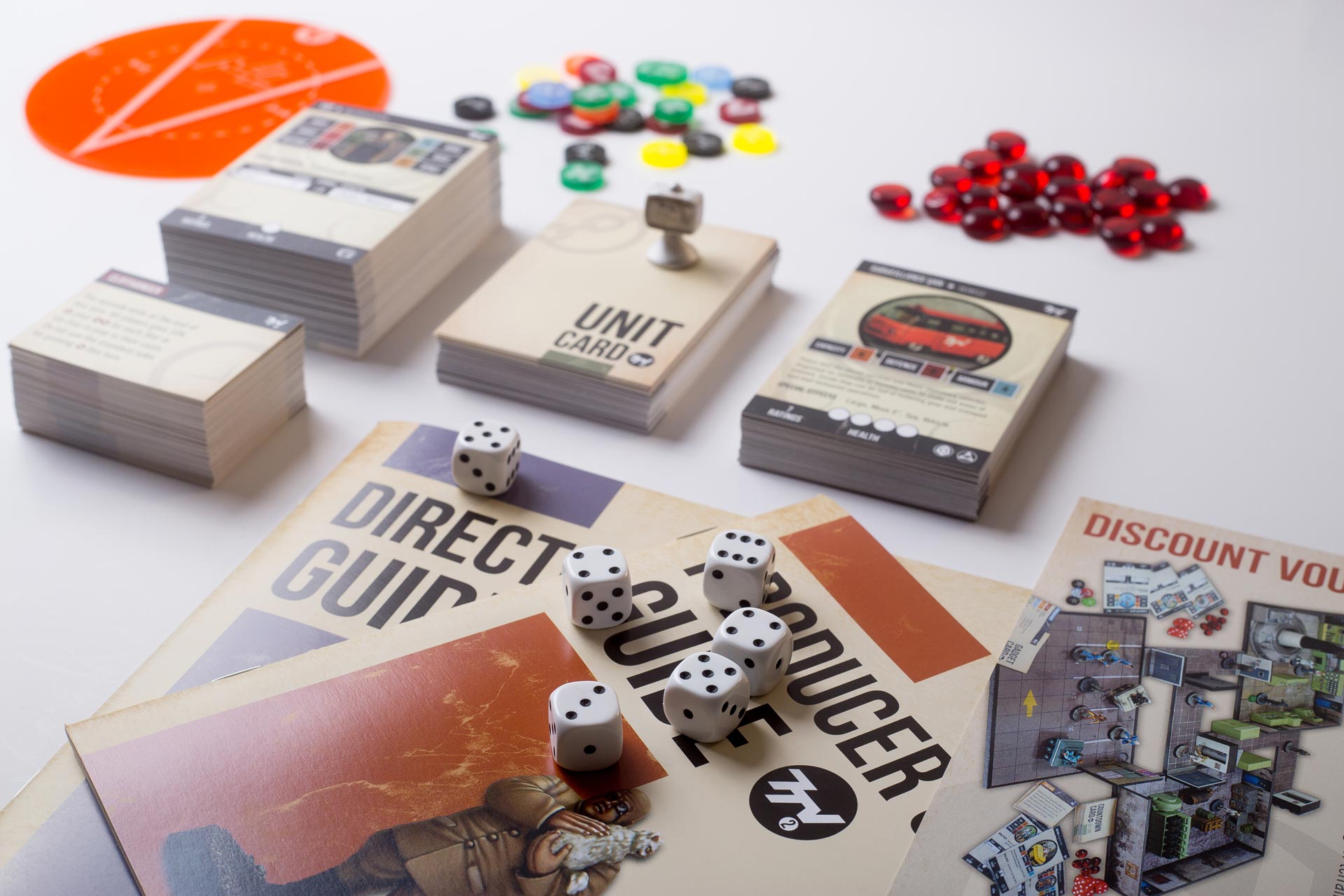 Tabletop miniatures game components including rulebooks, game boards, dice, coloured tokens, and red gemstone pieces